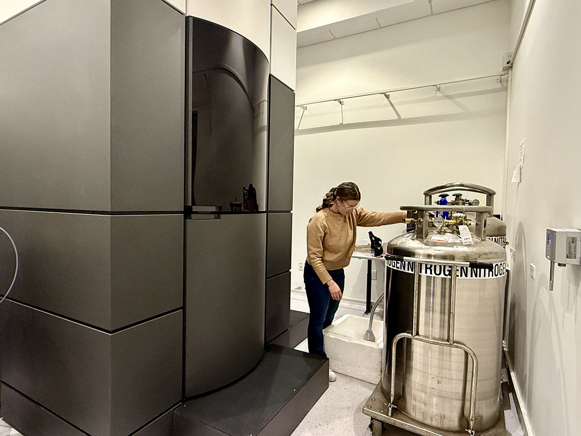 A Ph.D. student in a brown sweater stands next to a large steel tank